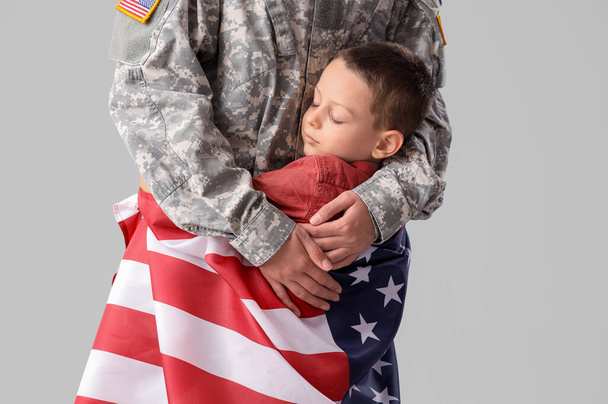 Female soldier hugging her little son with flag on light background. Veterans Day celebration